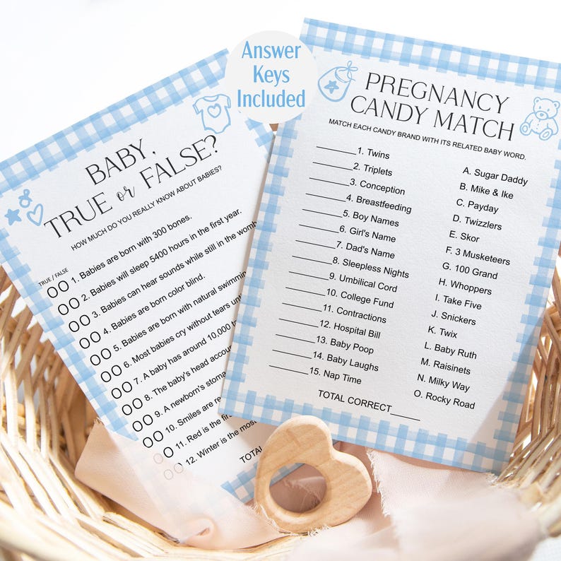 May include: Baby shower game cards with blue gingham borders. One card reads "Baby True or False?" and the other "Pregnancy Candy Match." A wooden heart-shaped teether is in the foreground. "Answer Keys Included" is printed on a circular sticker.