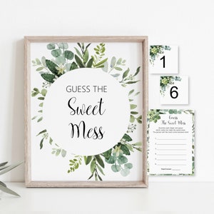 May include: A framed sign with the text "GUESS THE Sweet Mess" surrounded by a wreath of green leaves. Two small cards with the numbers 1 and 6, and a game card with instructions are also visible.