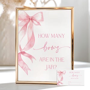 May include: A framed sign with a gold border features a watercolor illustration of pink bows and the text "HOW MANY bows ARE IN THE JAR?" A smaller matching card is also present. The sign is likely for a baby shower or party game.