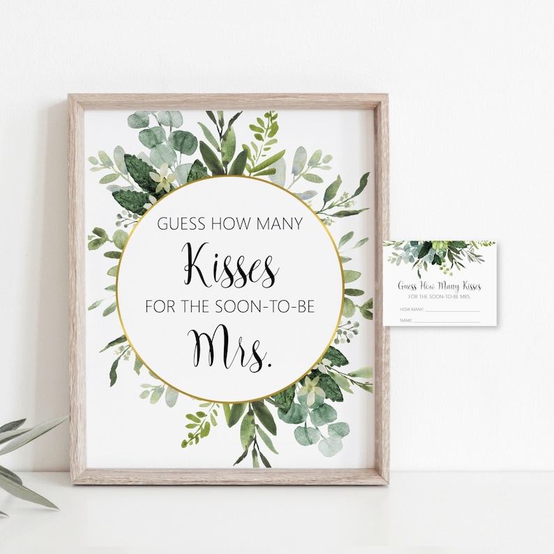 May include: A printable bridal shower game features a gold-framed greenery design.  The game asks guests to guess how many kisses the bride-to-be will receive.  A smaller matching card is included for guests to write their guesses and names. The main sign has a gold circular frame with the text 'Guess How Many Kisses for the Soon-to-be Mrs.' in elegant calligraphy. The greenery design is a watercolor style with eucalyptus and other leafy greens in varying shades of green.