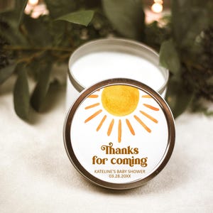 May include: A white candle in a silver tin with a yellow sun and orange rays graphic. The label reads "Thanks for coming" with the text "KATELINE'S BABY SHOWER 03.28.20XX" below.