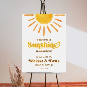 K&ouml;nnte beinhalten: Ein wei&szlig;es Willkommens-Schild mit einer gelben Aquarell-Sonnenillustration. Der Text lautet "a little ray of Sunshine is almost here! Welcome to Melissa & Ben's Baby Shower 06.10.2023"