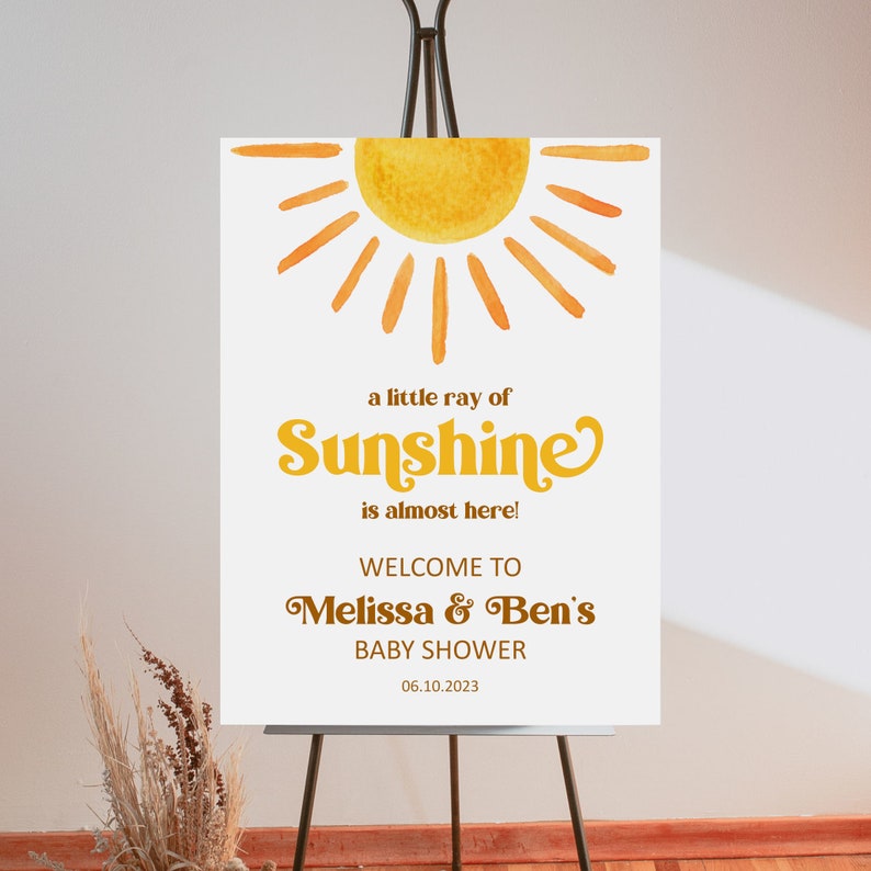 K&ouml;nnte beinhalten: Ein wei&szlig;es Willkommens-Schild mit einer gelben Aquarell-Sonnenillustration. Der Text lautet "a little ray of Sunshine is almost here! Welcome to Melissa & Ben's Baby Shower 06.10.2023"