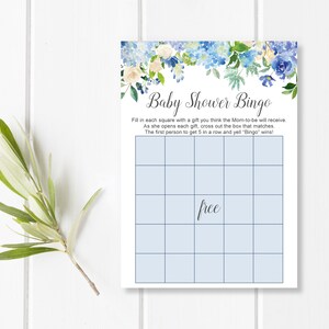 May include: Baby Shower Bingo card with a floral design in shades of blue and white. The card has a grid for playing the game, with the word "free" in the center. The text reads "Baby Shower Bingo" and instructions for the game.