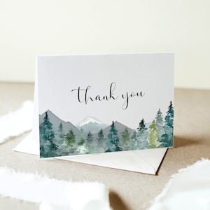 May include: A folded white card with a watercolor landscape design featuring mountains and evergreen trees in shades of green and blue. The words "Thank you" are written in elegant black script. An envelope is visible beneath the card.