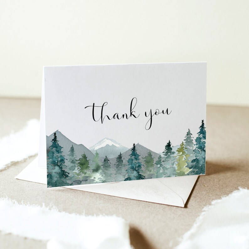 May include: A folded white card with a watercolor landscape design featuring mountains and evergreen trees in shades of green and blue. The words "Thank you" are written in elegant black script. An envelope is visible beneath the card.