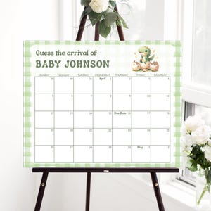 May include: A baby shower calendar on an easel, with a green and white gingham border. The text reads "Guess the arrival of BABY JOHNSON." A cartoon dinosaur and eggs are in the top right corner. The calendar grid is for April and May.