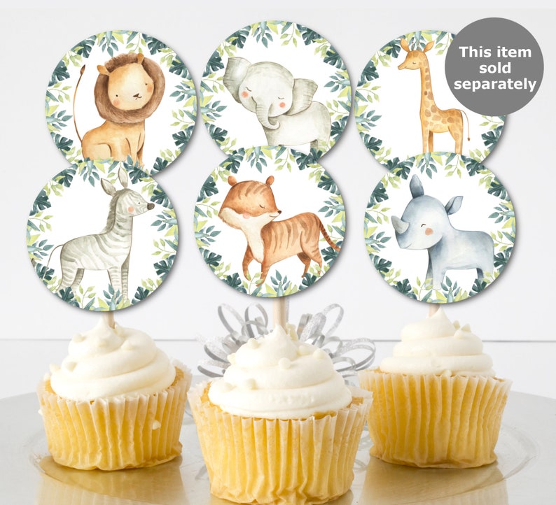 May include: Cupcakes with toppers featuring watercolor illustrations of safari animals, including a lion, elephant, giraffe, zebra, tiger, and rhino. The toppers are surrounded by a green leaf border. The cupcakes have white frosting.