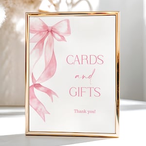 May include: A framed sign with a gold border. The sign features a watercolour illustration of a pink bow and ribbons. The words "CARDS and GIFTS" are written in pink script. The words "Thank you!" are written below.