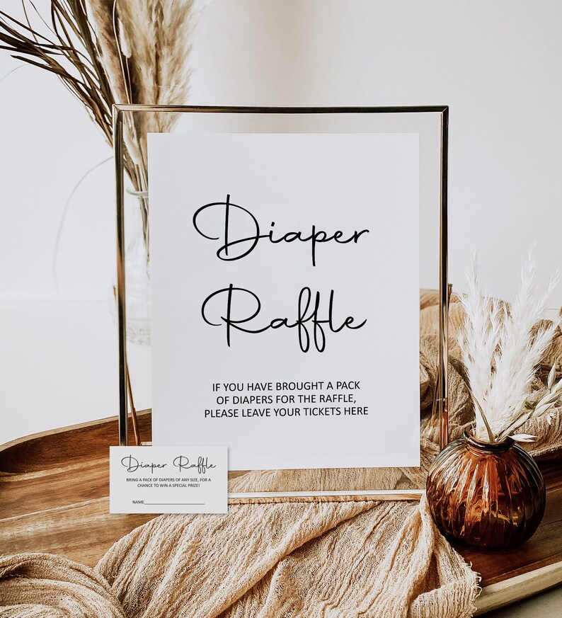 Puede incluir: Un cartel con marco dorado con el texto "Diaper Raffle" e instrucciones para que los invitados dejen sus boletos si trajeron pa&ntilde;ales. Una tarjeta peque&ntilde;a con el texto "Diaper Raffle" est&aacute; sobre una superficie de madera.