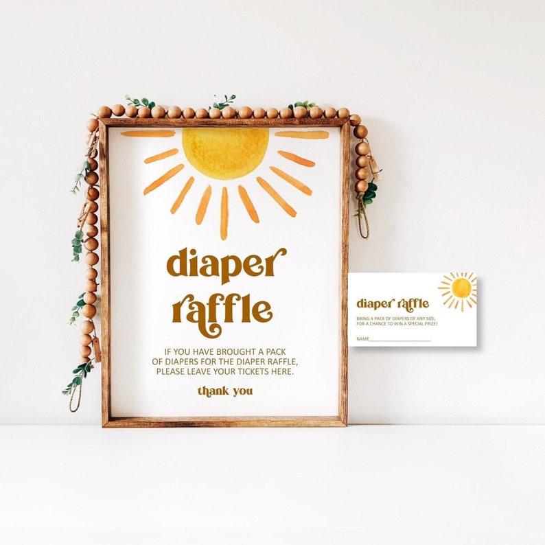 May include: A brown wooden frame with a white background and a yellow sun illustration. The text "diaper raffle" is written in brown letters. The text "If you have brought a pack of diapers for the diaper raffle, please leave your tickets here. Thank you" is written in black letters. A smaller white card with a yellow sun illustration and the text "diaper raffle" is also visible.