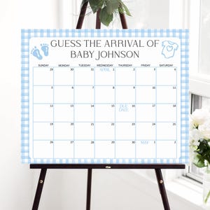 May include: A baby shower calendar on an easel, featuring a blue gingham border. The calendar reads "Guess the Arrival of Baby Johnson" and includes a calendar grid with the due date marked on April 15th.