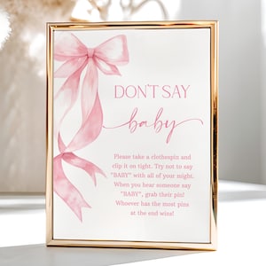May include: A framed sign with a gold border features the text "DON'T SAY baby" in pink. A watercolor-style pink bow and ribbons adorn the left side. The sign's text describes a baby shower game. The background is white.