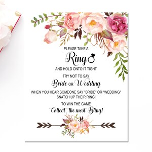 May include: A white card with pink and green floral illustrations. The text reads: "Please take a Ring and hold onto it tight. Try not to say Bride or Wedding. When you hear someone say "Bride" or "Wedding" snatch up their ring! To win the game collect the most Bling!"