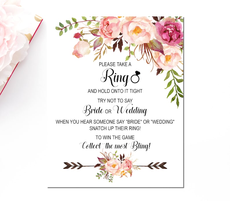 May include: A white card with pink and green floral illustrations. The text reads: "Please take a Ring and hold onto it tight. Try not to say Bride or Wedding. When you hear someone say "Bride" or "Wedding" snatch up their ring! To win the game collect the most Bling!"