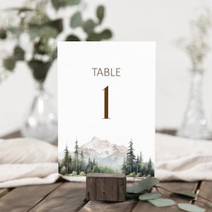 May include: A white table number card with the word "TABLE" and the number "1" in brown. The card features a watercolor landscape of mountains and trees. The card is held by a small wooden block.