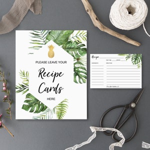 May include: A white sign with the text "PLEASE LEAVE YOUR Recipe Cards HERE" surrounded by green tropical leaves and a gold pineapple. A recipe card and black scissors are also visible.