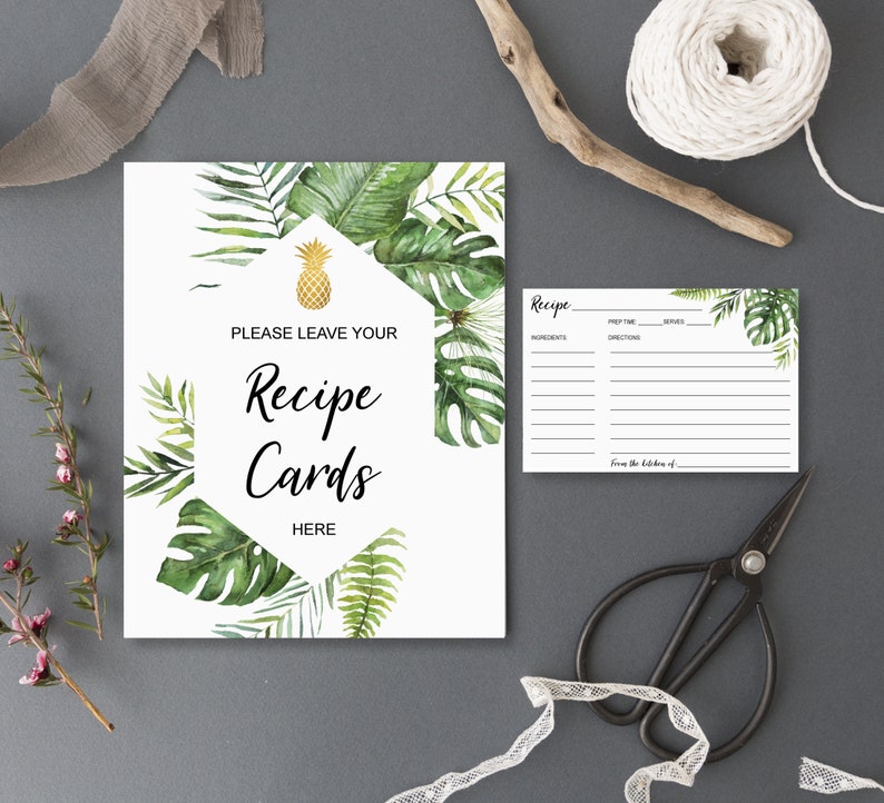 May include: A white sign with the text "PLEASE LEAVE YOUR Recipe Cards HERE" surrounded by green tropical leaves and a gold pineapple. A recipe card and black scissors are also visible.