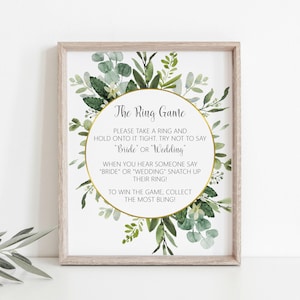 Peut inclure: Une affiche imprimable avec une bordure florale verte et du texte dor&eacute; qui dit "The Ring Game". Les instructions du jeu sont de prendre un anneau et de le tenir fermement, en essayant de ne pas dire "Bride" ou "Wedding". Lorsque quelqu'un dit "Bride" ou "Wedding", attrapez son anneau. Pour gagner le jeu, collectez le plus de bling!