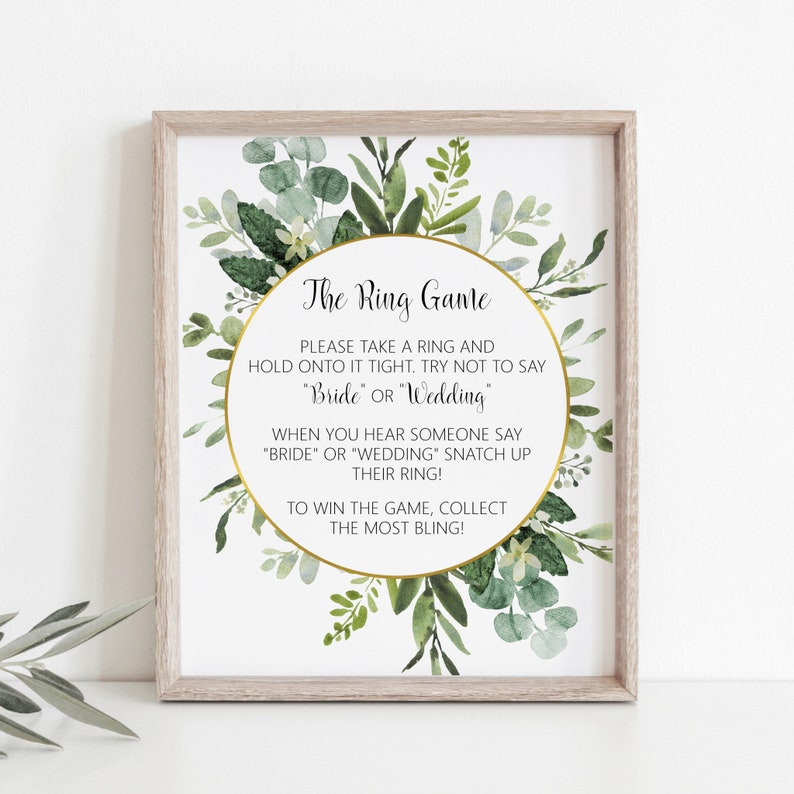 Peut inclure: Une affiche imprimable avec une bordure florale verte et du texte dor&eacute; qui dit "The Ring Game". Les instructions du jeu sont de prendre un anneau et de le tenir fermement, en essayant de ne pas dire "Bride" ou "Wedding". Lorsque quelqu'un dit "Bride" ou "Wedding", attrapez son anneau. Pour gagner le jeu, collectez le plus de bling!