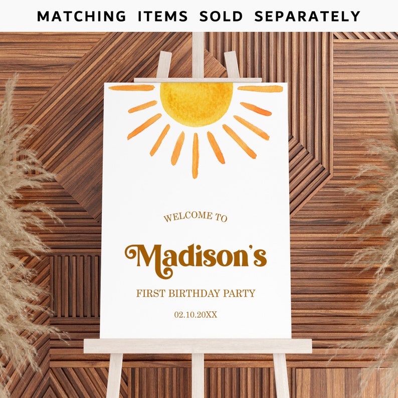 May include: Welcome to Madison's First Birthday Party sign with a watercolor sun illustration. The sign is on a wooden easel and has a white background with brown text.
