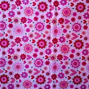 May include: A pink fabric with a repeating pattern of flowers and hearts in shades of pink and red. The flowers have multiple layers of petals and are surrounded by dotted circles. Hearts and small flowers fill the spaces.