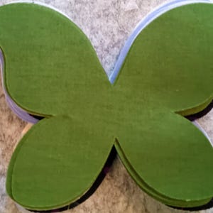 May include: A stack of green butterfly-shaped cutouts. The cutouts are made of a textured material and are layered on top of each other. The edges of the cutouts are a lighter green, and the stack is on a neutral background.