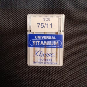May include: A pack of three gold sewing needles in a clear plastic case with a blue label that reads "Universal Titanium Klasse 125 Years German Experience" and "Size 75/11".
