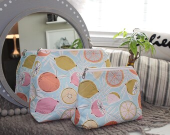 May include: Three quilted cosmetic bags with a light blue background and a pattern of citrus fruits in pink, yellow, and orange. The bags have silver zippers and are arranged in a group, varying in size.