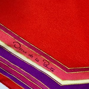 May include: A silk scarf with a vibrant red background and a geometric border design in shades of purple, pink, and gold. The scarf features the signature "Oscar de la Renta" in cursive script.