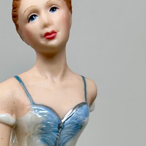 Könnte beinhalten: Eine Porzellanfigur einer Ballerina, die ein weißes Tutu mit einem blauen Oberteil und einer silbernen Krone trägt. Die Ballerina ist in einer anmutigen Tanzpose mit ausgestreckten Armen dargestellt.