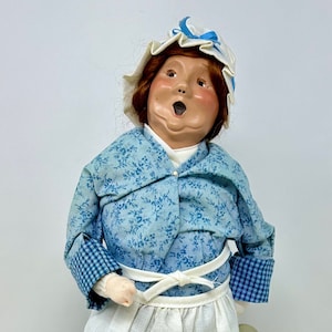May include: A vintage-style doll wearing a blue and white floral dress with a white apron and a white bonnet with a blue ribbon. The doll has a rosy complexion and is singing with her mouth open.