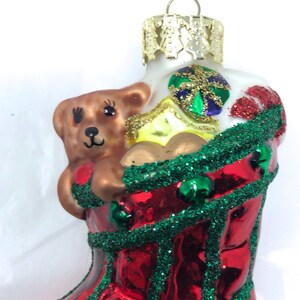 May include: Red and green glass Christmas bauble shaped like a stocking with a brown teddy bear inside. The stocking is decorated with glitter and green beads.
