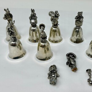 May include: A collection of silver bells with various animal figurines on top. The bells are arranged on a white surface. Some of the figurines include a rabbit, a squirrel, a cat, and a bird.