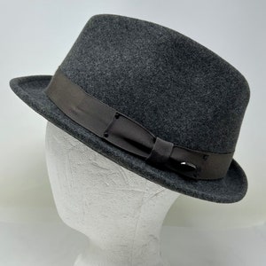 Pode incluir: Um chapéu fedora de feltro cinza com uma fita marrom e uma fivela. O chapéu está em uma cabeça de manequim branca.