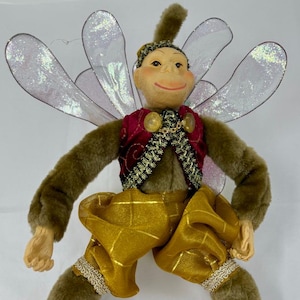 May include: A whimsical fairy doll with a smiling face, brown fur arms and legs, and iridescent wings. The doll wears a burgundy vest with gold accents, gold patterned pants, and red shoes. A decorative item.