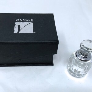 May include: A clear glass bottle with a silver cap and a faceted design. The bottle is in a black box with the word "VANMARK" printed on the top.