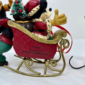 May include: A Christmas ornament featuring Mickey Mouse dressed as Santa Claus in a red suit and hat, riding in a red sleigh pulled by a brown and green figure. The ornament is made of painted resin and has a gold metal hanger. The ornament is labeled "&copy; DISNEY" and "DESIGNED BY MIDWEST OF CANNON FALLS".