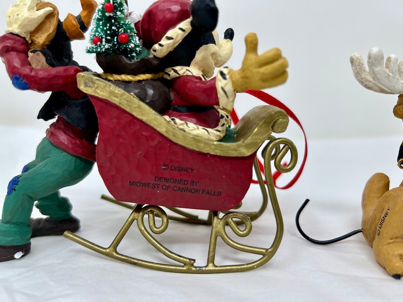 May include: A Christmas ornament featuring Mickey Mouse dressed as Santa Claus in a red suit and hat, riding in a red sleigh pulled by a brown and green figure. The ornament is made of painted resin and has a gold metal hanger. The ornament is labeled "&copy; DISNEY" and "DESIGNED BY MIDWEST OF CANNON FALLS".