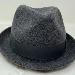 Pode incluir: Um chap&eacute;u fedora cinza-carv&atilde;o com um acabamento texturizado semelhante a feltro. O chap&eacute;u apresenta uma copa estruturada com um vinco central e frente pin&ccedil;ada, complementado por uma larga faixa de fita gorgor&atilde;o preta s&oacute;lida na base.