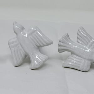 May include: Four white ceramic bird figurines in flight, arranged horizontally. Each bird has outstretched wings and detailed feathering. The birds are on a white surface, creating a clean and simple aesthetic.