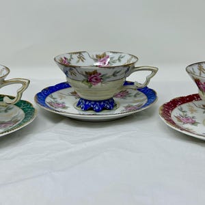May include: Set of three floral teacups and saucers. Each cup has a different colored base and features a floral design with pink roses and green leaves. The saucers have a matching floral design with gold accents.
