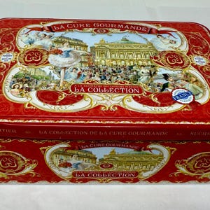 May include: A rectangular red tin box with ornate gold and white detailing. The lid features a detailed illustration of a building and people, with the text "LA CURE GOURMANDE" and "LA COLLECTION". The box is labeled "France".