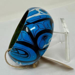 May include: A blue and black patterned bangle bracelet with a Native American design. The bracelet has a glossy finish and is displayed on a clear stand. The text "Angel L. Tingley" is printed on the inside of the bracelet.