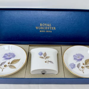 Puede incluir: Juego de porcelana Royal Worcester Bone China en una caja azul. El juego incluye dos platos ovalados pequeños y un recipiente rectangular, todos blancos con ribete dorado y un diseño floral en morado y marrón. La tapa de la caja tiene el nombre de la marca.
