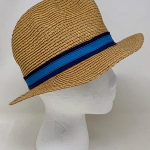 May include: A tan straw fedora hat featuring a textured woven pattern and a decorative band around the crown. The band consists of a central bright blue stripe flanked by two thinner dark navy blue stripes, creating a distinct color contrast.