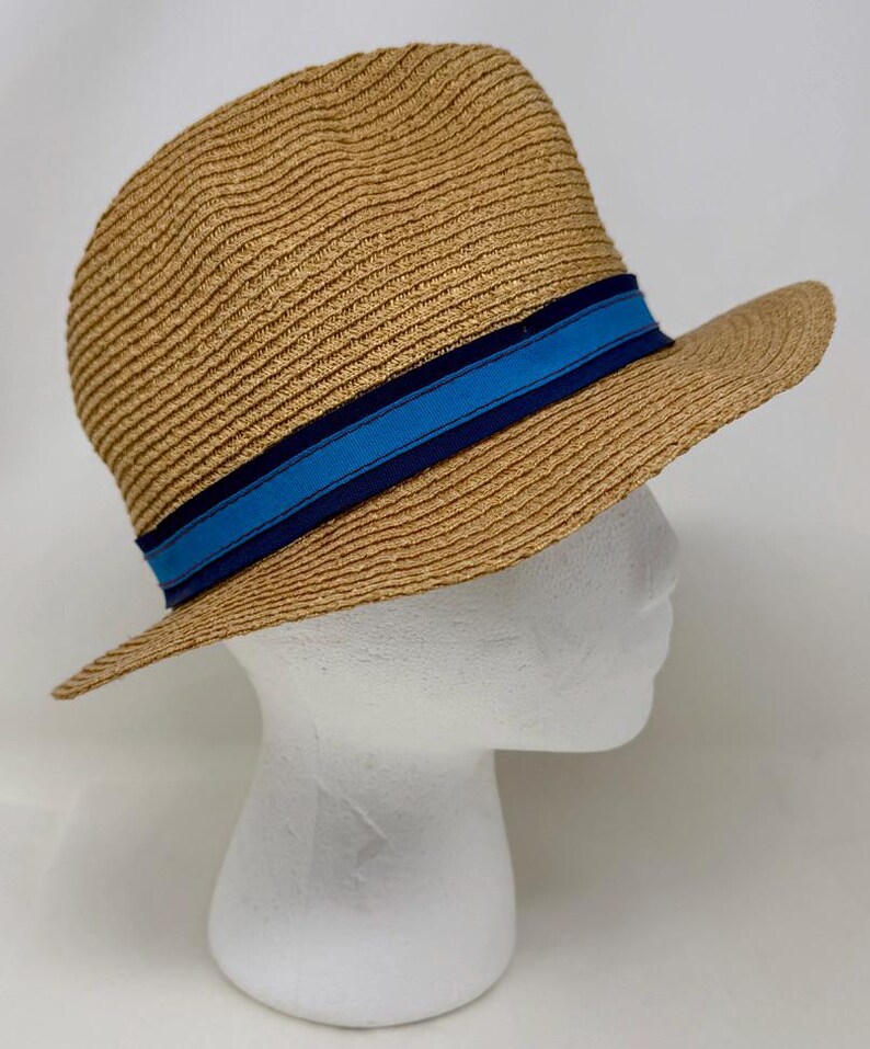 May include: A tan straw fedora hat featuring a textured woven pattern and a decorative band around the crown. The band consists of a central bright blue stripe flanked by two thinner dark navy blue stripes, creating a distinct color contrast.