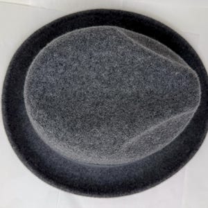 Pode incluir: Um chap&eacute;u fedora de feltro cinza-escuro visto diretamente de cima, exibindo a copa arredondada com um vinco central e uma aba circular larga. O material tem uma apar&ecirc;ncia macia e texturizada, apresentado contra um fundo liso.