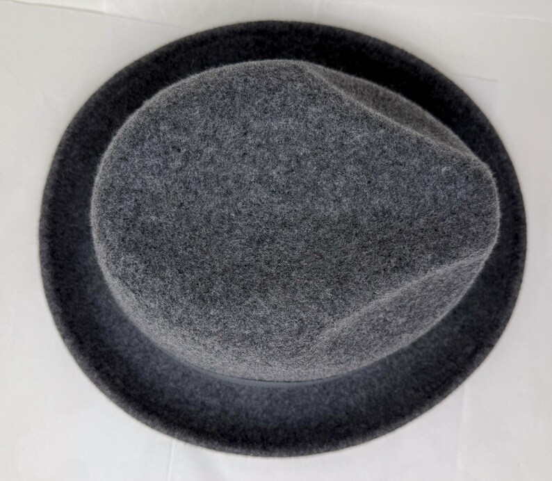 Pode incluir: Um chap&eacute;u fedora de feltro cinza-escuro visto diretamente de cima, exibindo a copa arredondada com um vinco central e uma aba circular larga. O material tem uma apar&ecirc;ncia macia e texturizada, apresentado contra um fundo liso.
