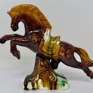 May include: A glossy ceramic horse figurine in a rearing pose, finished in a deep brown glaze with accents of green and yellow on the saddle and base. The sculpture features a textured mane and tail, with a stylized support pillar underneath.